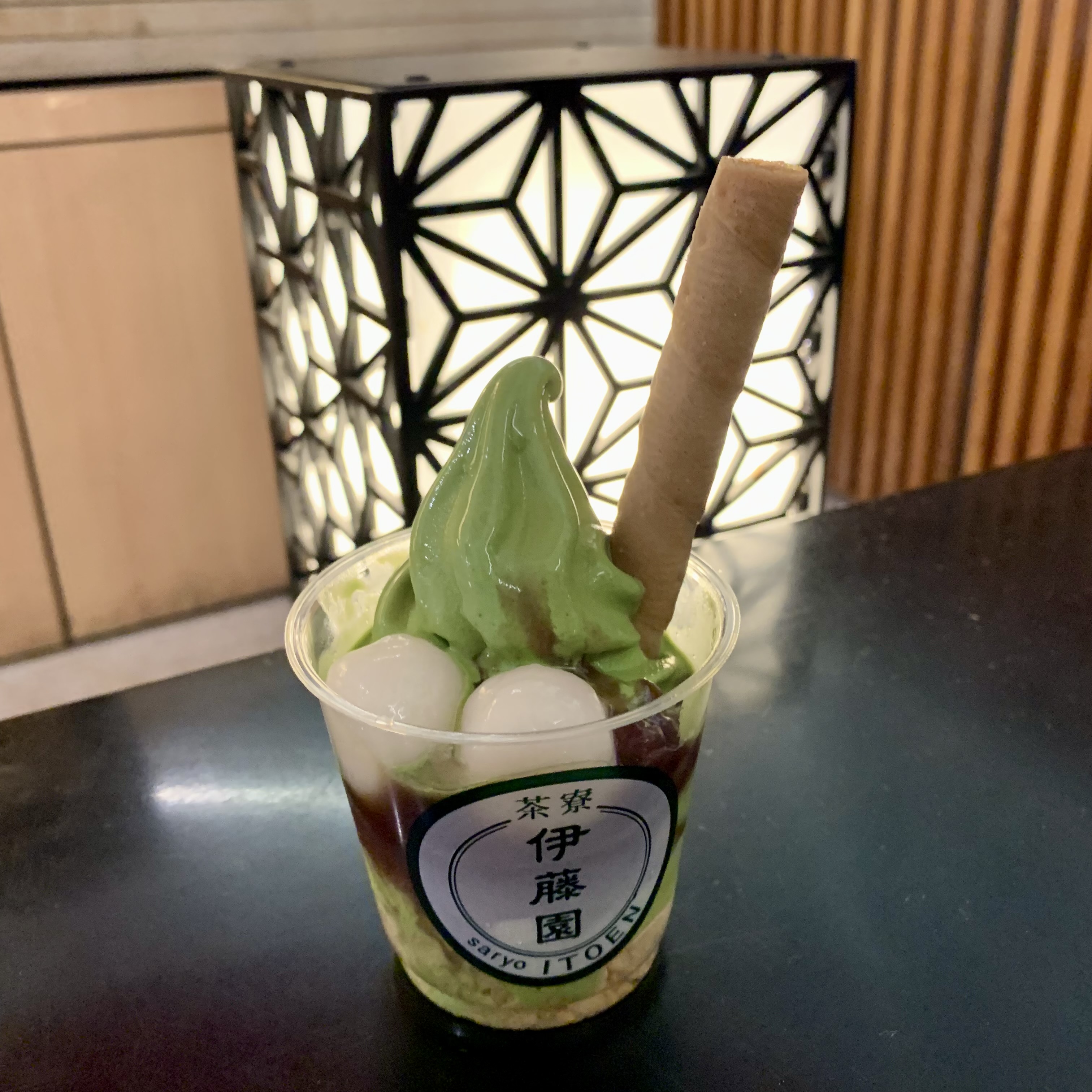 画像には、「伊藤園」と書かれたカップに入った抹茶ソフトクリームのデザートが映っています。デザートには、抹茶ソフトクリームの上に白玉団子が2つトッピングされ、さらにウエハースのような細長いクッキーが立てられています。背景には幾何学模様の明かりが見え、デザートが室内で撮影されたことがわかります。全体的に、日本の抹茶を使ったデザートの美味しさと雰囲気が感じられます。